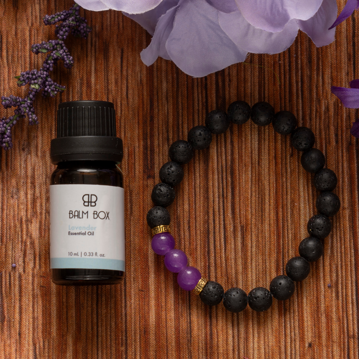 A bottle labeled Premium Soothing Skin & Lips sits next to a beaded bracelet with black and purple beads on a brown striped surface surrounded by purple flowers—an ideal self-care gift for cancer patients.