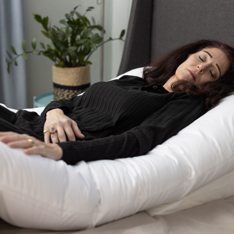 A woman with dark hair lies on a bed, eyes closed, hugging the Sleep Again Pillow System—a pillow designed for side sleepers. A potted plant is visible in the background.