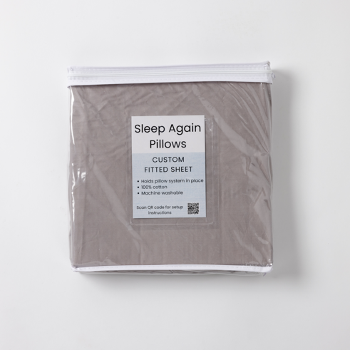 The Custom Fitted Sheet for Sleep Again Pillow System is a packaged gray, 100% cotton, machine washable sheet that helps with pillow positioning and includes a QR code for setup instructions.