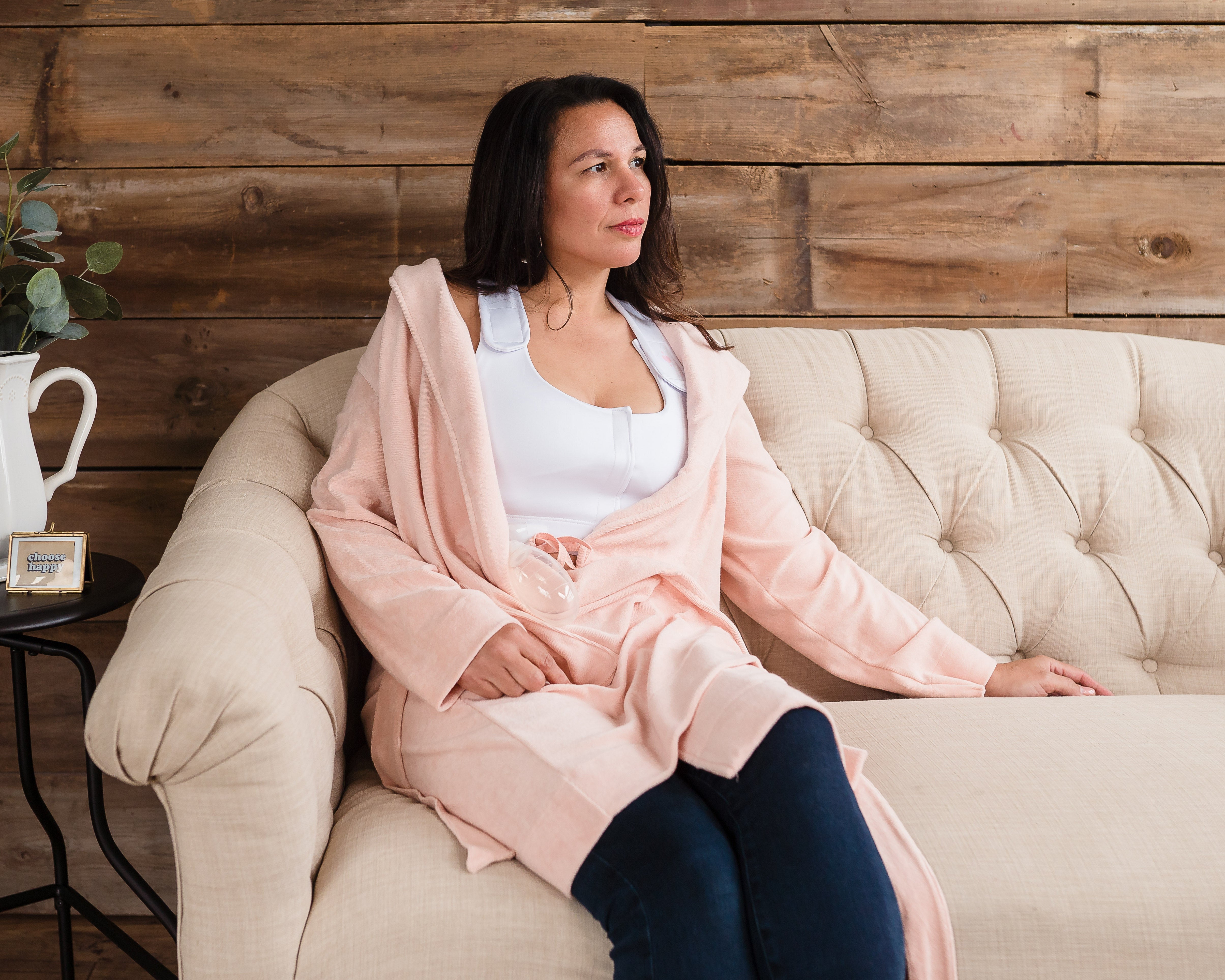 A woman in a pale pink robe and white top, possibly wearing the Larissa Post-Surgical Bra for breast surgery recovery, sits on a cream tufted sofa. On a nearby round black table are a white pitcher with greenery and a small box.