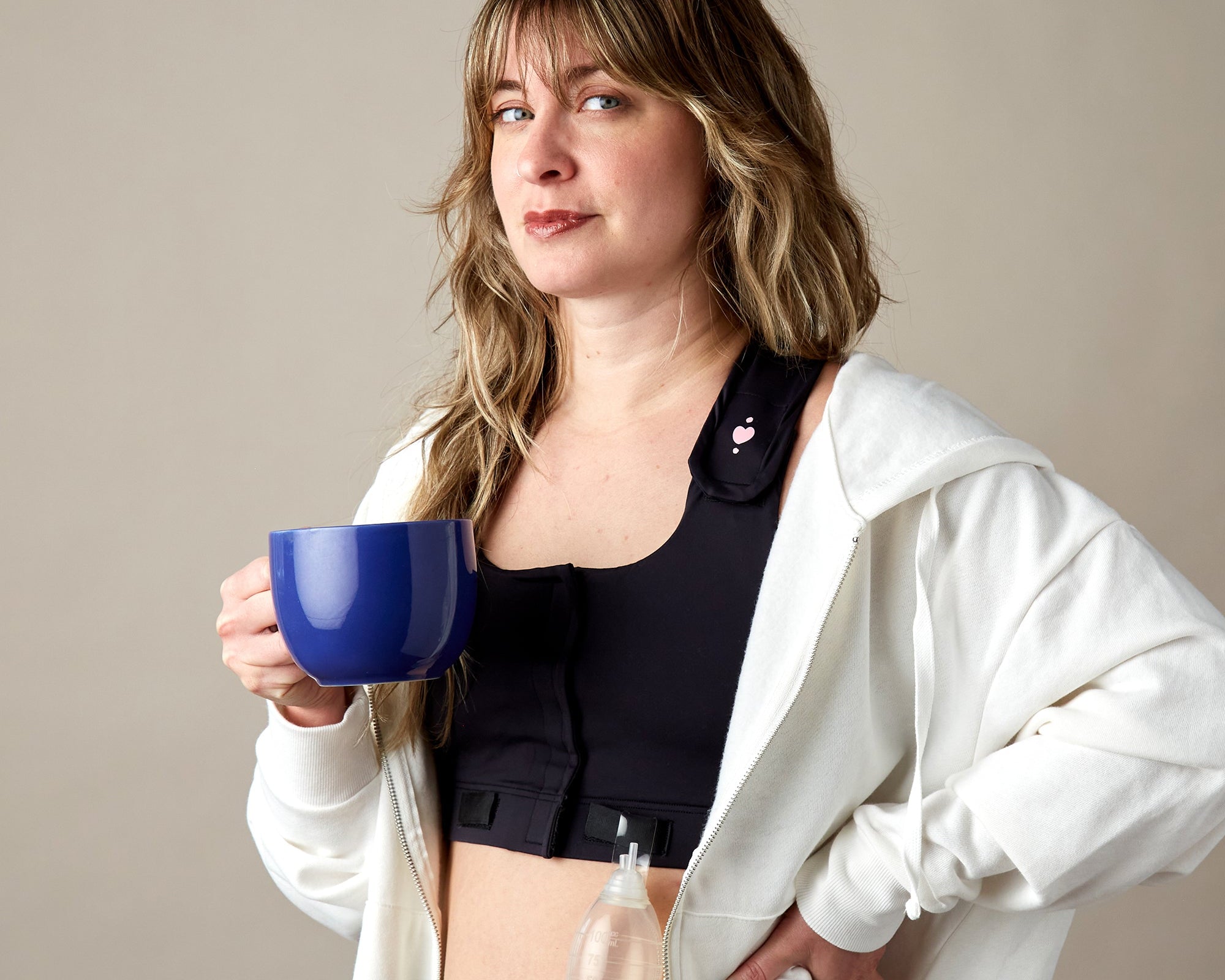 A woman with wavy hair wears a white hoodie over the Larissa Post-Surgical Bra, holding a blue mug. A surgical drain and drainage tube are attached, illustrating breast surgery recovery against a plain beige background.