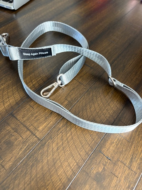 A gray adjustable Sleep Again Surgical Drain Holder with metal clips and a black Sleep Again Pillows label is shown on a wooden surface, perfect for managing surgical drains after surgery.