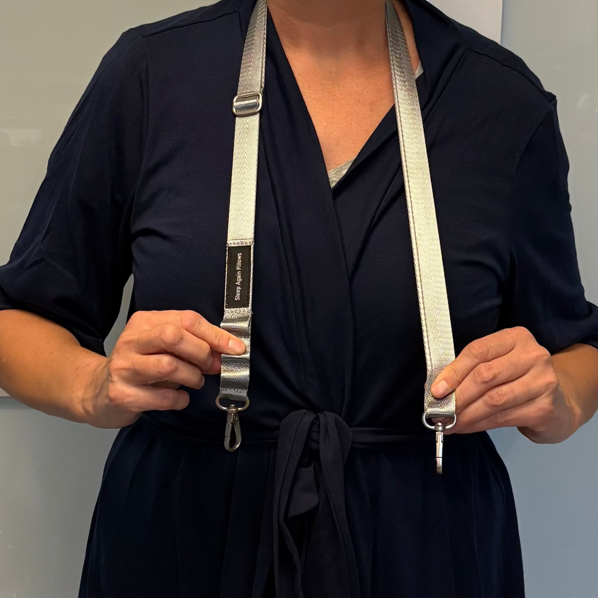 A person in a navy blue wrap dress holds the Sleep Again Surgical Drain Holder, featuring a metallic, seatbelt-style adjustable strap, clips at each end, and a black label by the shoulder for post-op drain management.
