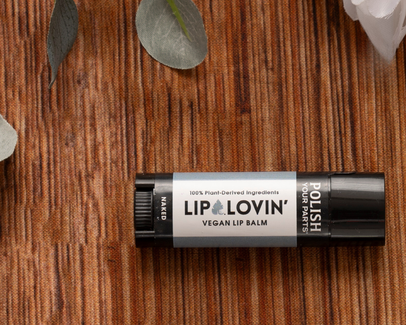 A black tube of Premium Soothing Skin & Lips balm rests on a brown textured surface amid eucalyptus and white flowers—ideal for soothing lips or adding to a cancer care gift box. The label states: “100% Plant-Derived Ingredients.”.