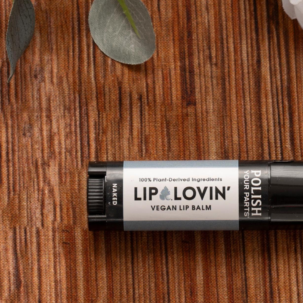 A black tube of Premium Soothing Skin & Lips balm rests on a brown textured surface amid eucalyptus and white flowers—ideal for soothing lips or adding to a cancer care gift box. The label states: “100% Plant-Derived Ingredients.”.