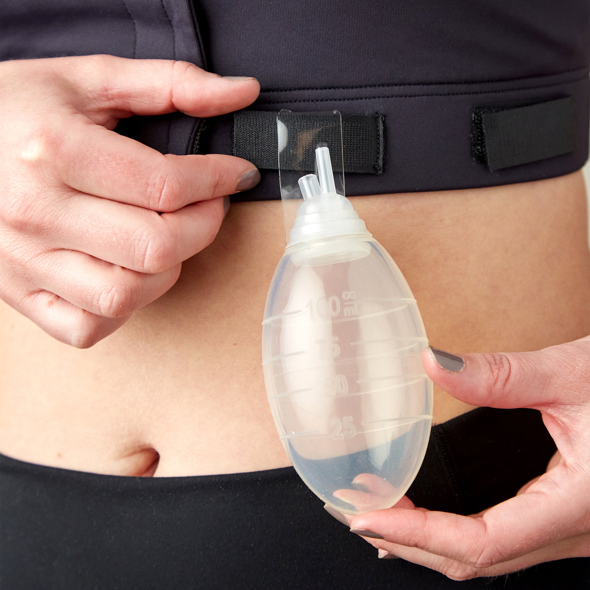Using the Larissa Post Surgical Bra, a person attaches a medical drain bulb to the waistband for effective drain management. One hand holds the bulb while the other secures it with a Velcro strap—ideal for mastectomy recovery.