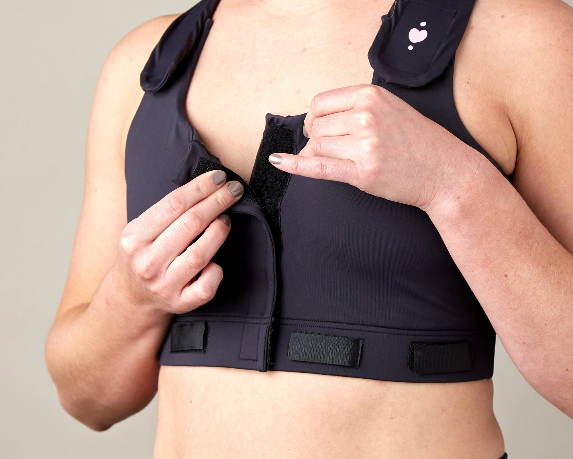 A person fastens the front of the Larissa Post-Surgical Bra in black, using hook-and-loop closures to show its adjustable design, ideal for breast surgery recovery.