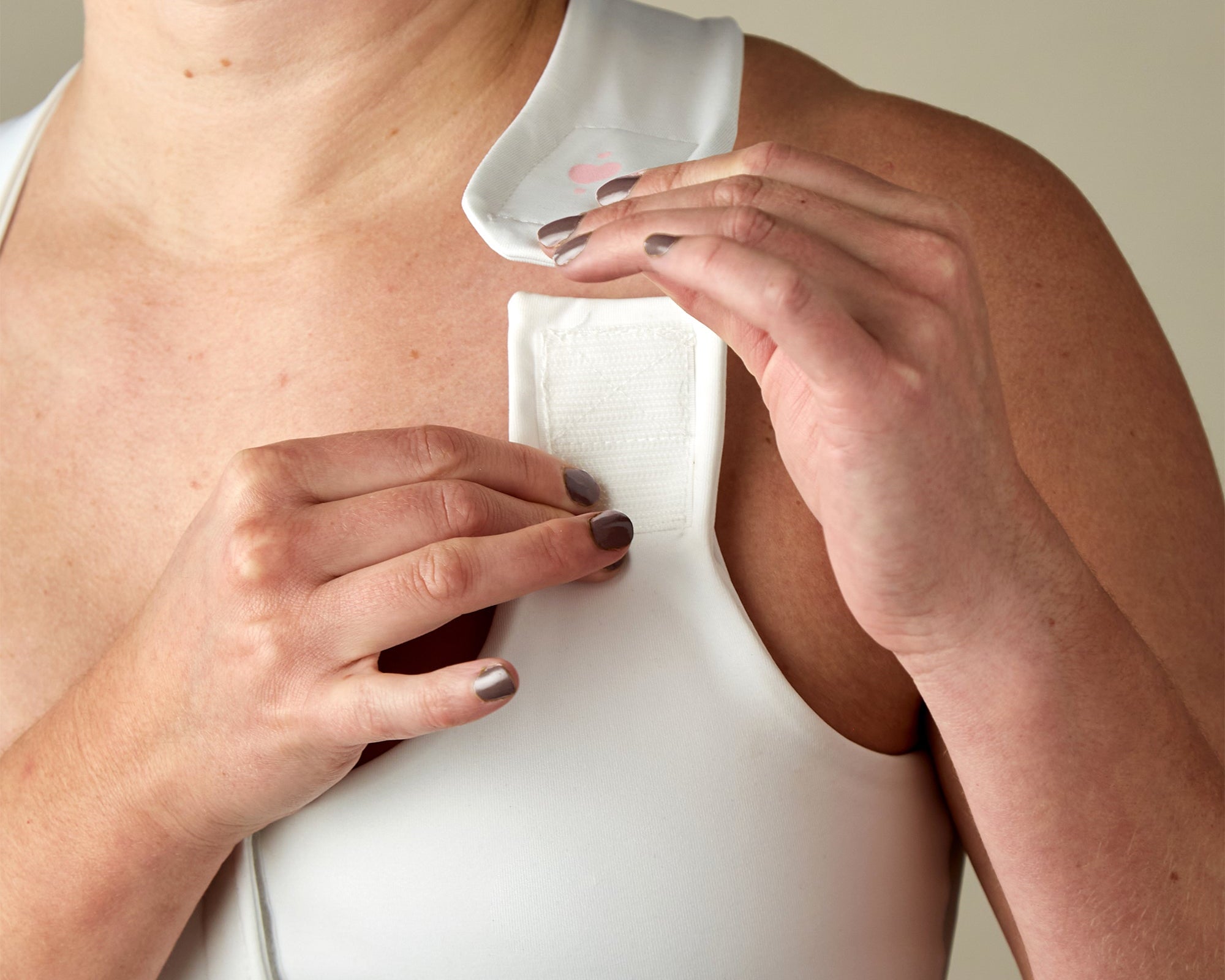 A person with light skin fastens the Velcro shoulder strap of a Larissa Post-Surgical Bra in white. Only the upper chest, shoulder, and hands are visible against a neutral background, highlighting breast surgery recovery.