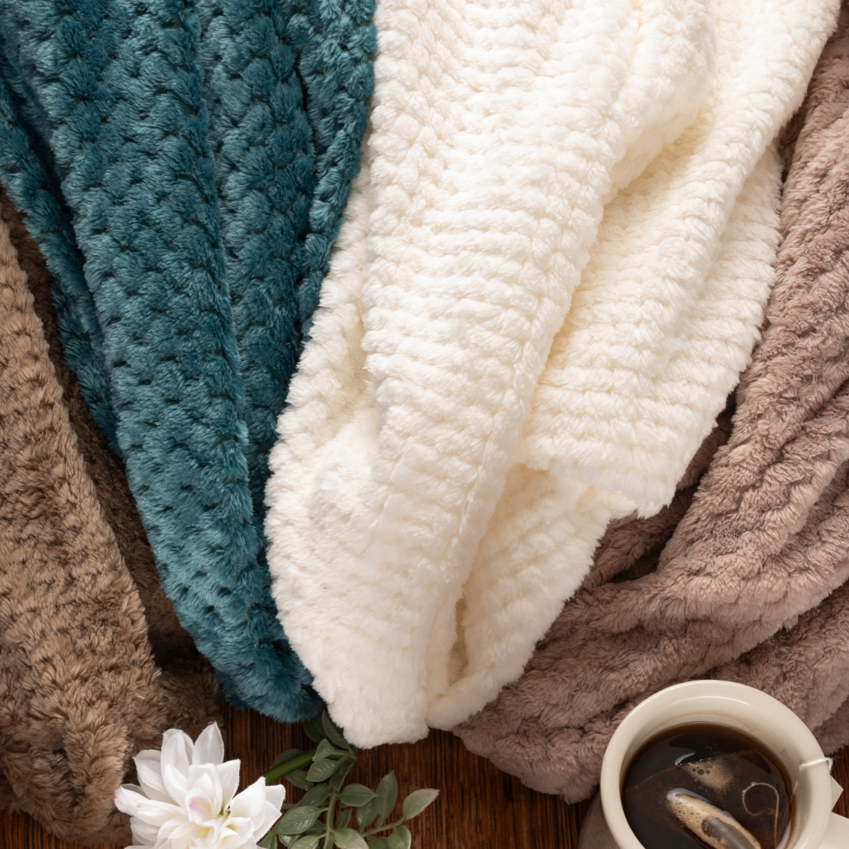 Four soft, textured blankets in brown, teal, white, and taupe are draped beside a white mug of tea and flower—offering cozy comfort found in the Sweetest Rest & Recovery Box, perfect for any cancer recovery gift.
