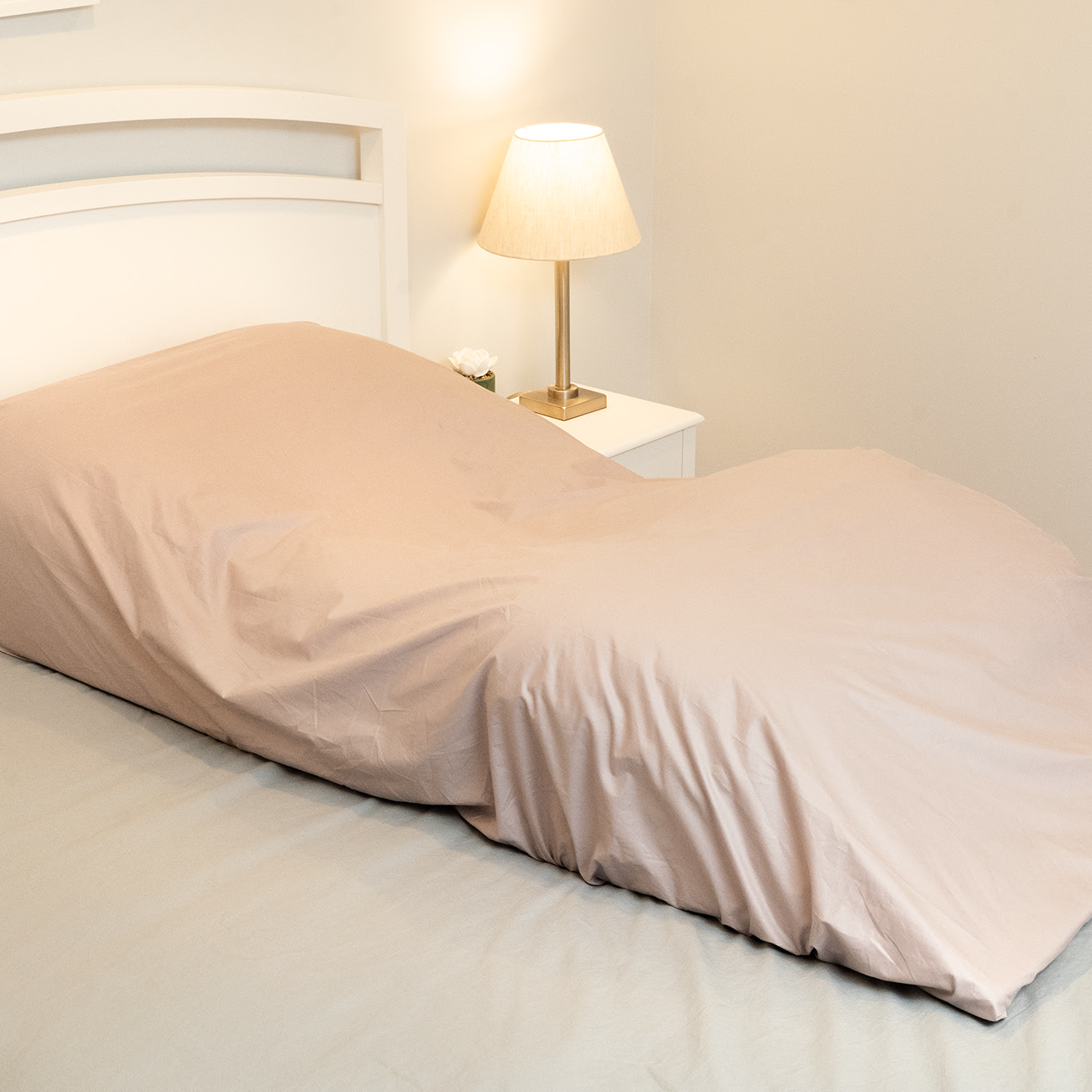 A bed features the Custom Fitted Sheet for Sleep Again Pillow System over a human-shaped pillow, resembling a person lying down; a nightstand with lamp and plant adds to the thoughtful presentation.