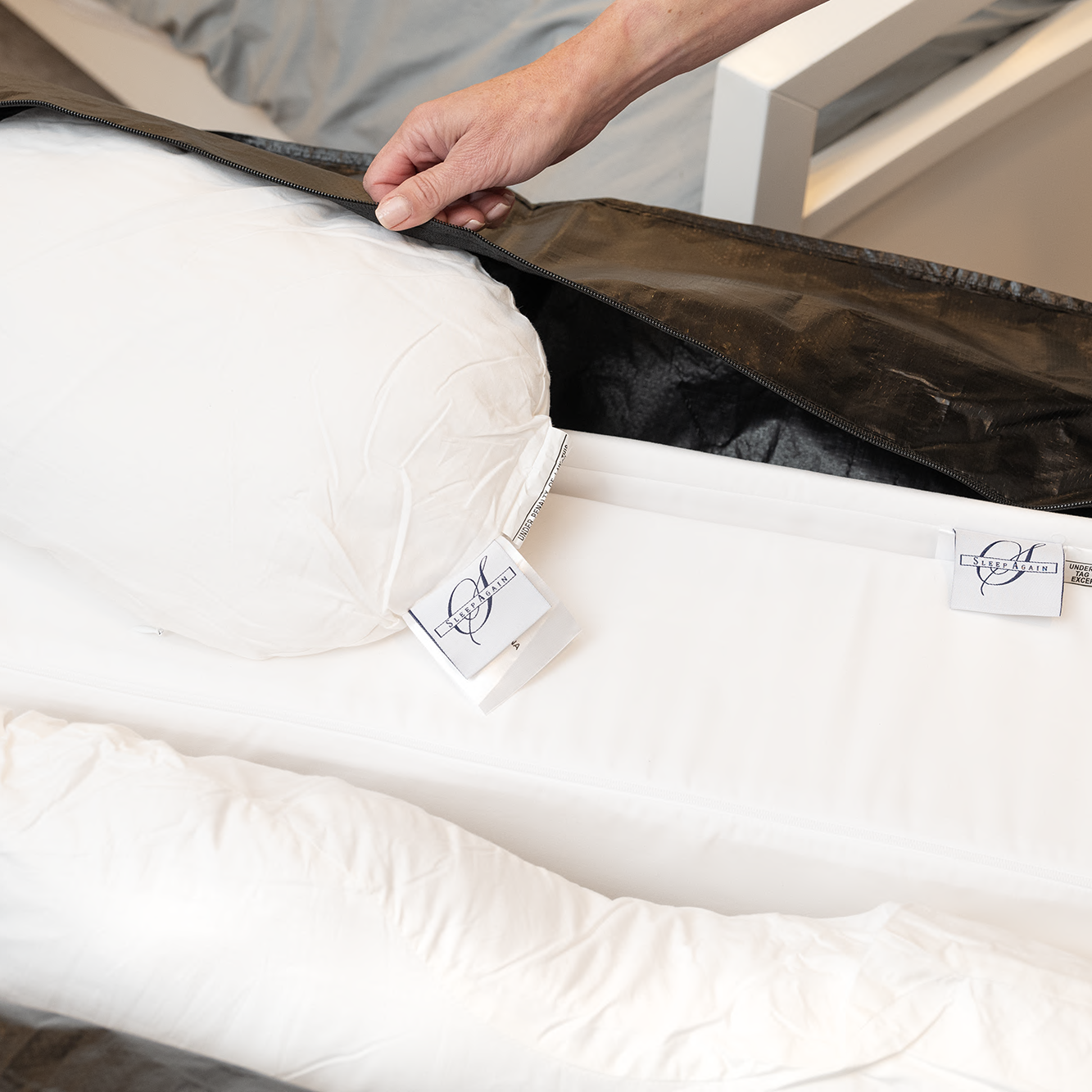 A hand lifts the black Travel and Storage Bag for Sleep Again Pillow System, revealing a white body pillow with tags inside, water resistant and ideal for travel, next to a bed with gray sheets in the background.