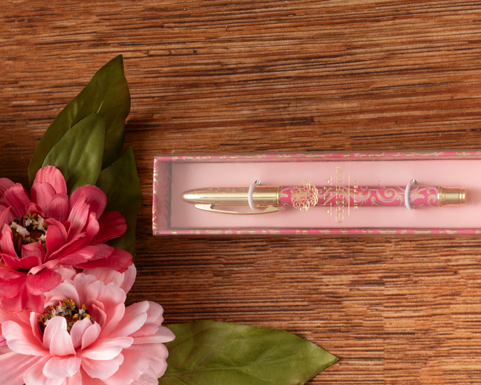The Sweetest Rest & Recovery Box, featuring a decorative pink and gold pen, is displayed on a wooden surface beside pink artificial flowers with green leaves.