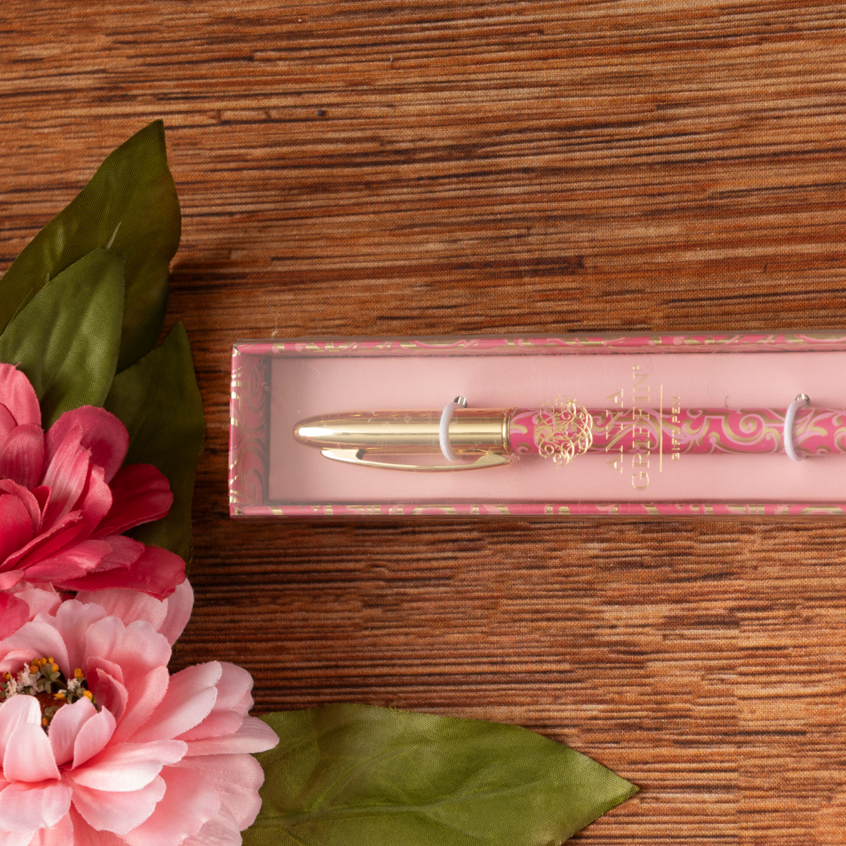 The Sweetest Rest & Recovery Box, featuring a decorative pink and gold pen, is displayed on a wooden surface beside pink artificial flowers with green leaves.
