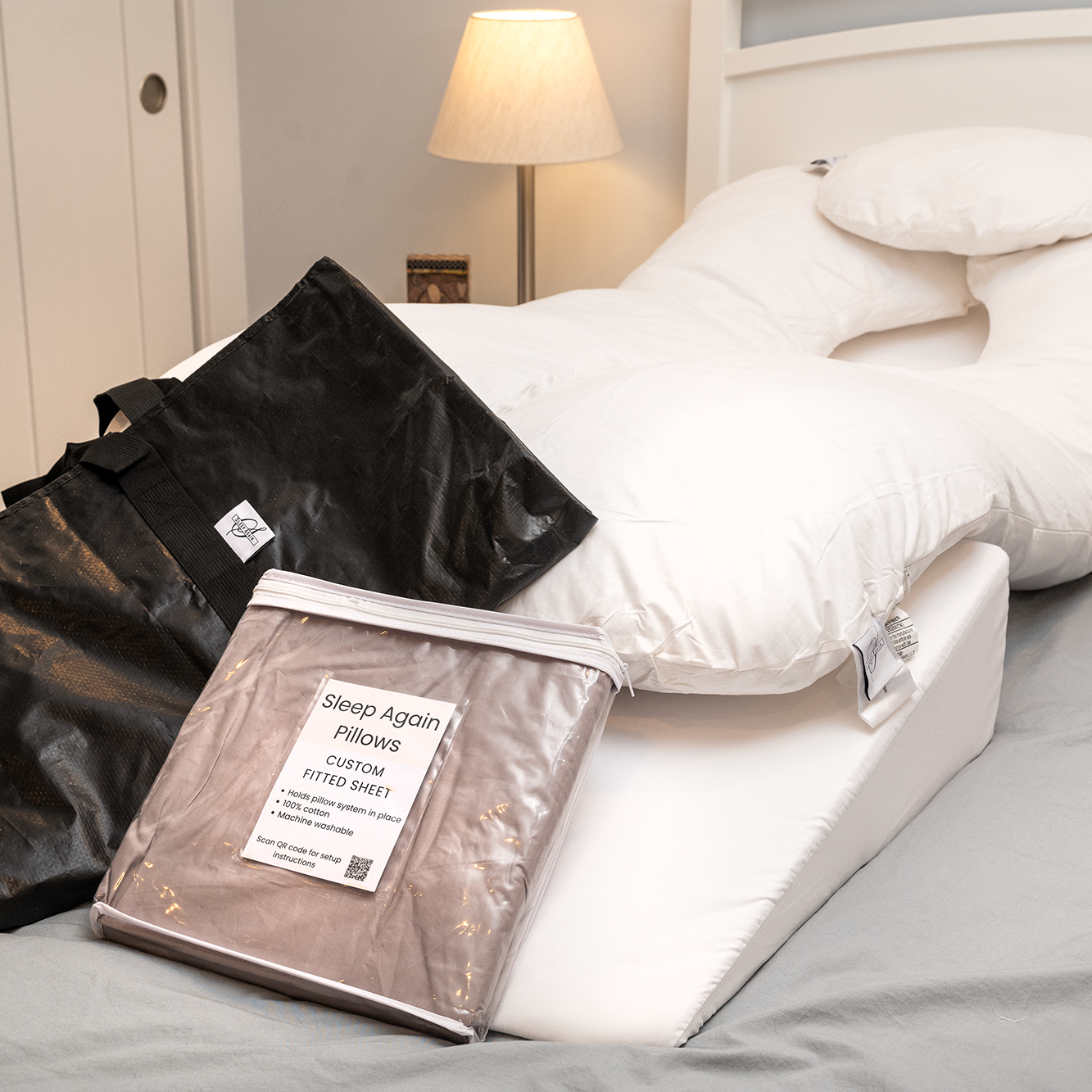 A neatly made bed features the Sleep Again Pillow System Bundle, white bedding, a head pillow, a bedside lamp, and two product bags labeled Sleep Again Pillow System and custom fitted sheet on top.