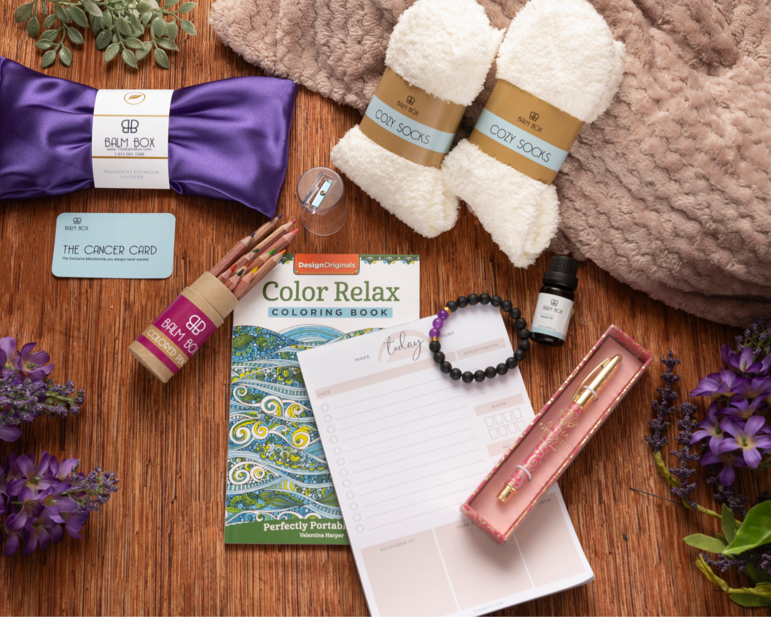 The Sweetest Rest & Recovery Box is a cozy cancer recovery gift featuring a purple sleep mask, fuzzy socks, bath soak, planner, coloring book, pen set, bracelet, essential oil, gift card, and purple flowers on a soft brown blanket.