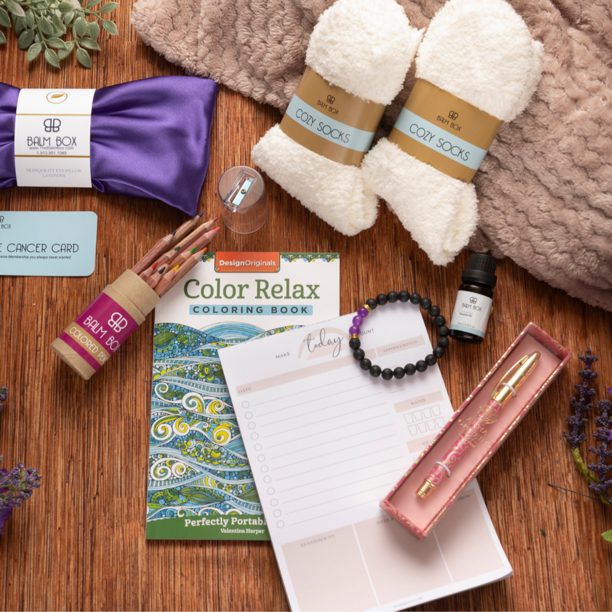 The Sweetest Rest & Recovery Box is a cozy cancer recovery gift featuring a purple sleep mask, fuzzy socks, bath soak, planner, coloring book, pen set, bracelet, essential oil, gift card, and purple flowers on a soft brown blanket.