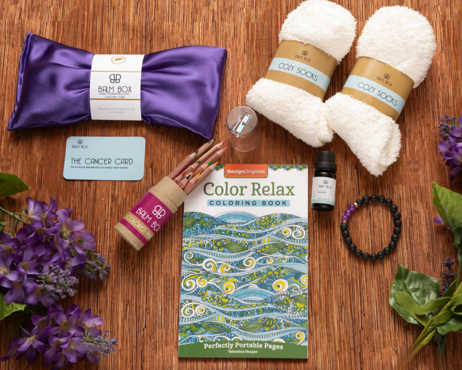 The Sweetest Rest & Recovery Box on a wooden surface includes cozy socks, a lavender eye pillow, an inspirational coloring book, colored pencils, essential oil, a bracelet, lip balm, a Cancer Card, and purple flowers.