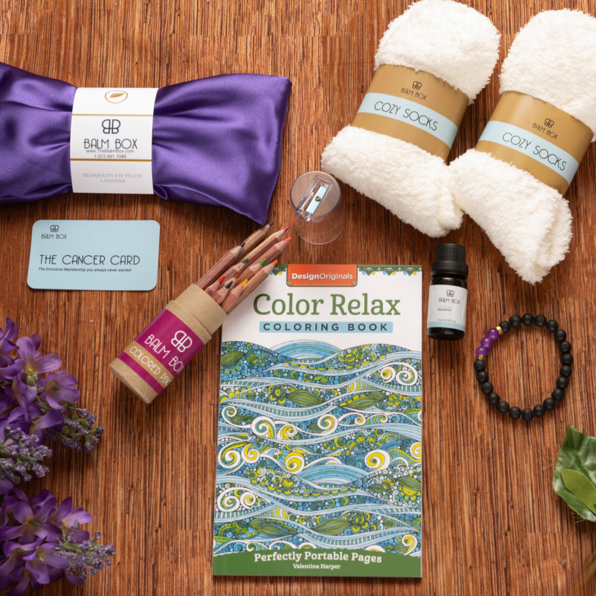 The Sweetest Rest & Recovery Box on a wooden surface includes cozy socks, a lavender eye pillow, an inspirational coloring book, colored pencils, essential oil, a bracelet, lip balm, a Cancer Card, and purple flowers.