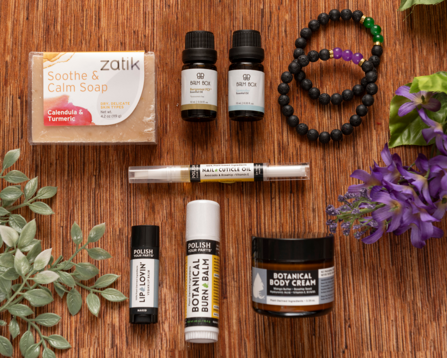 A flat lay of Premium Soothing Skin & Lips shows soap, essential oils, a cuticle oil pen, lip and burn balms, body cream, beaded bracelets, and flowers—a thoughtful self-care gift for cancer patients.