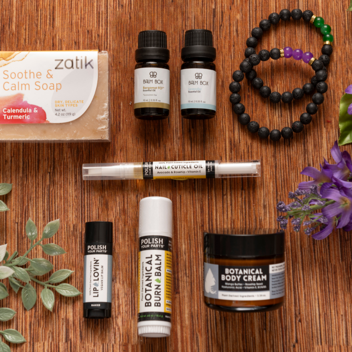 A flat lay of Premium Soothing Skin & Lips shows soap, essential oils, a cuticle oil pen, lip and burn balms, body cream, beaded bracelets, and flowers—a thoughtful self-care gift for cancer patients.