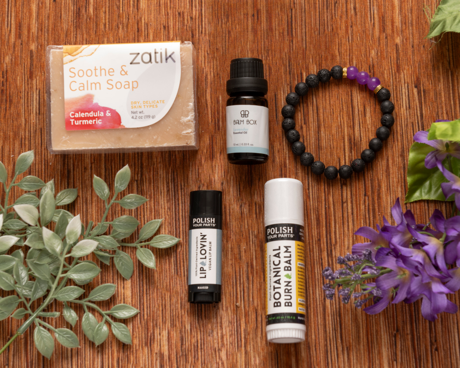 Premium Soothing Skin & Lips: a flat lay set of soap, essential oil, two lip products, and a black bracelet with purple beads, accented by a purple flower—ideal for soothing care or as a thoughtful cancer support gift.