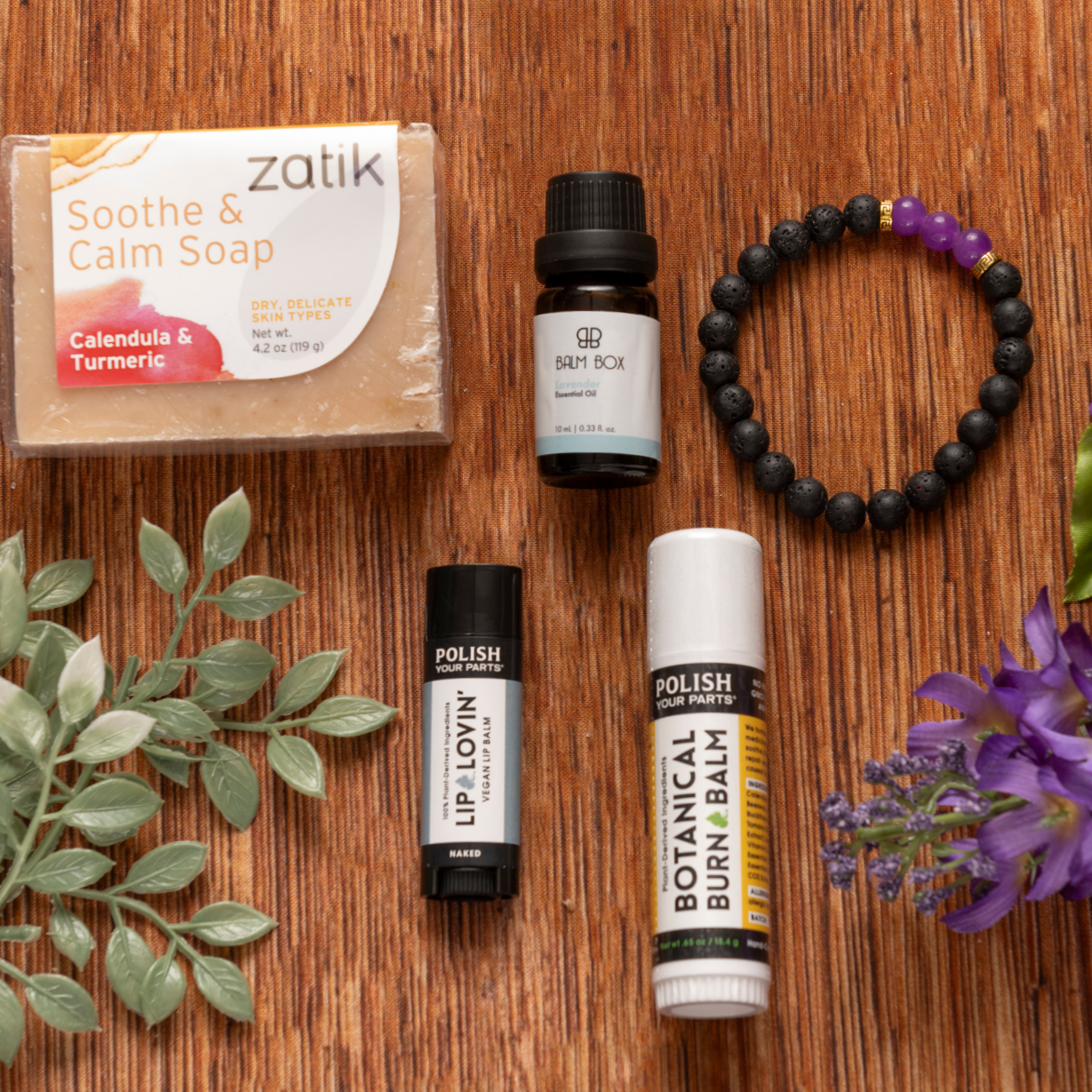 Premium Soothing Skin & Lips: a flat lay set of soap, essential oil, two lip products, and a black bracelet with purple beads, accented by a purple flower—ideal for soothing care or as a thoughtful cancer support gift.