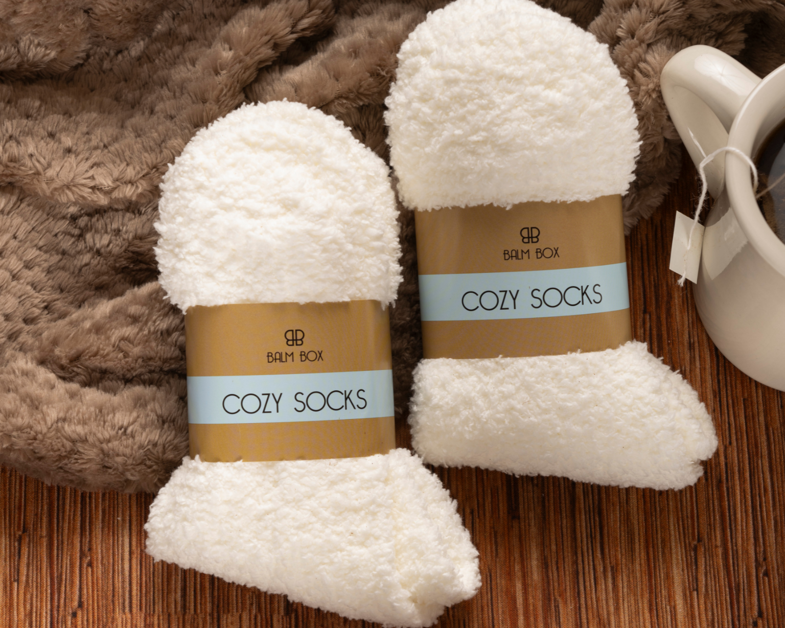 Two pairs of “Cozy Socks” lie on a wooden surface with a fuzzy brown blanket and white tea mug—perfect comforts included in The Best Mastectomy Care Package for a thoughtful breast cancer gift.