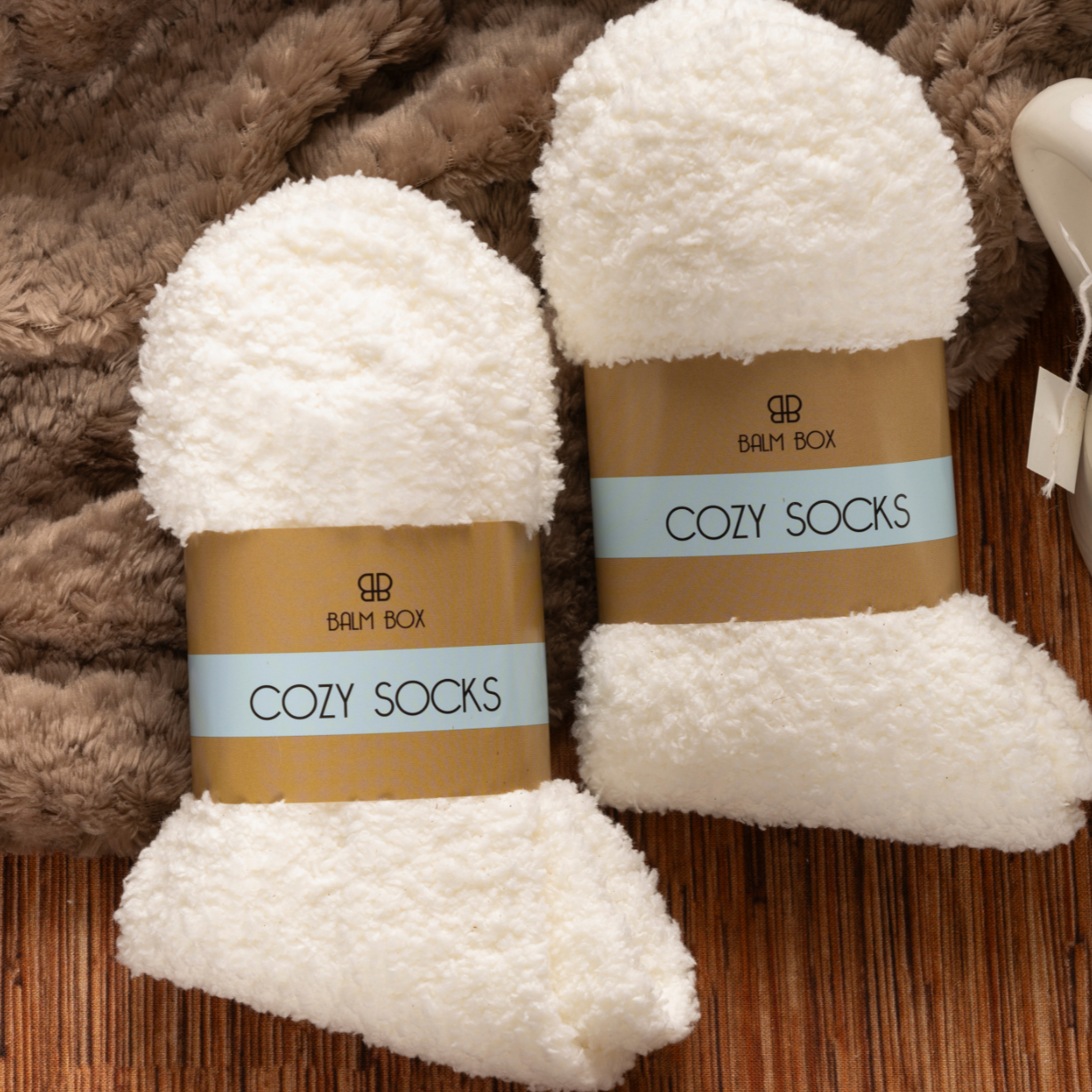 Two pairs of “Cozy Socks” lie on a wooden surface with a fuzzy brown blanket and white tea mug—perfect comforts included in The Best Mastectomy Care Package for a thoughtful breast cancer gift.