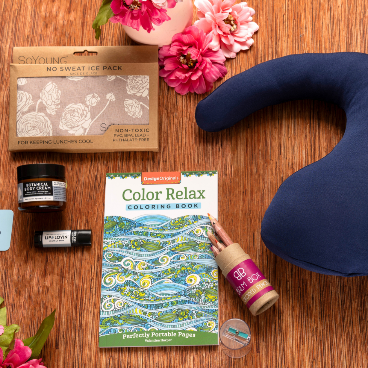 A flat lay of The Best Mastectomy Care Package—featuring a post-mastectomy pillow, ice pack, lip balm, hand sanitizer, coloring book, pen, “The Cancer Card,” and artificial flowers—makes an ideal breast cancer gift box on a wooden surface.