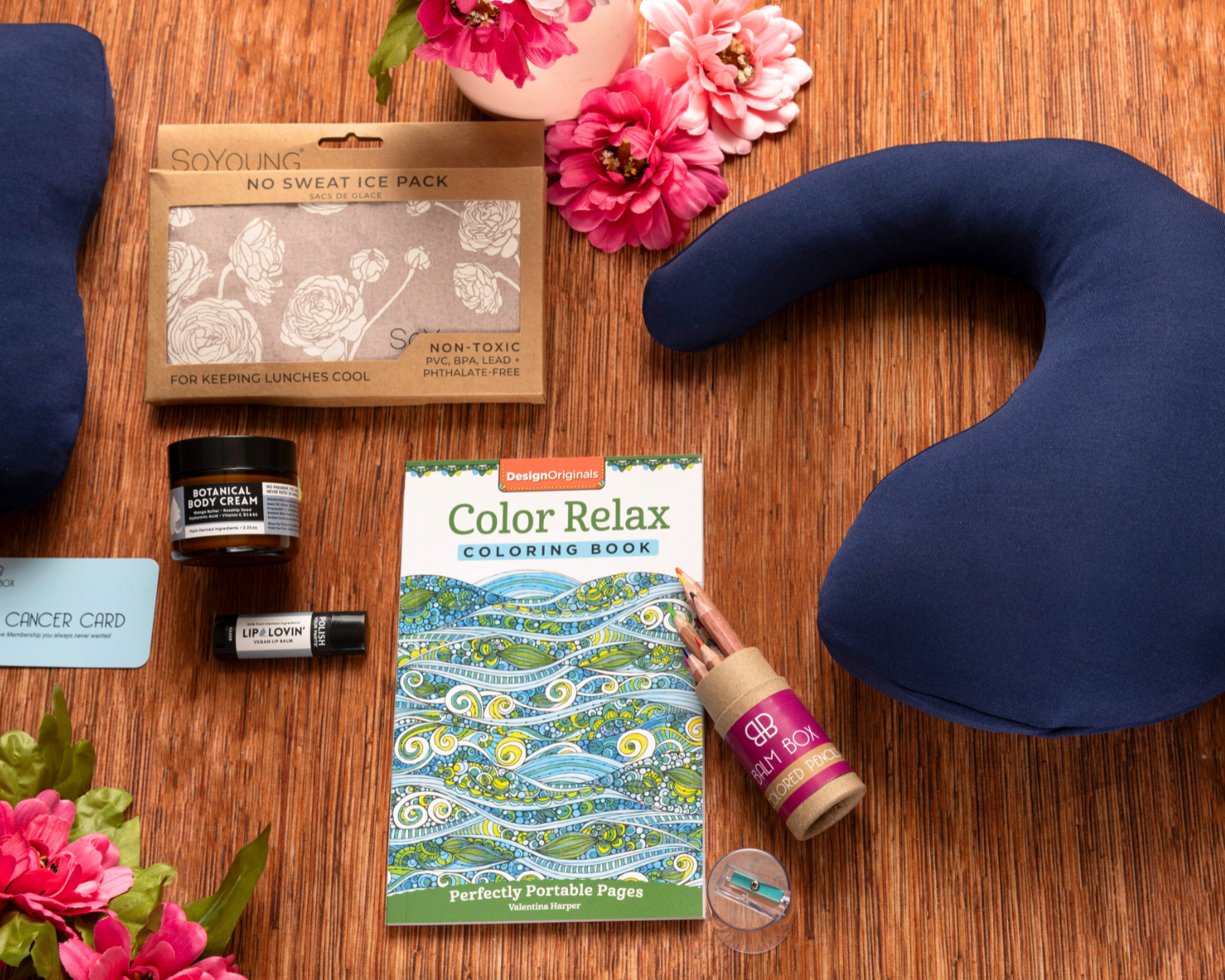 The Best Mastectomy Care Package on a wooden surface features a blue neck pillow, eye mask, coloring book, lip balm, lotion, chill pack, gift card, and pink flowers—ideal for comfort and support after a mastectomy.