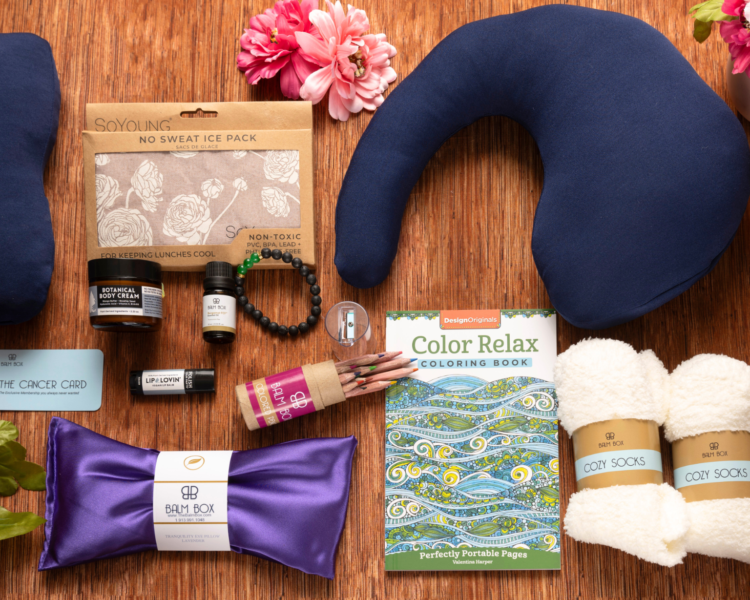 The Best Mastectomy Care Package displayed on a wooden surface includes a neck pillow, socks, coloring book, eye mask, ice pack, small jars of cream, colored pencils, and decorative pink flowers—an ideal breast cancer gift.
