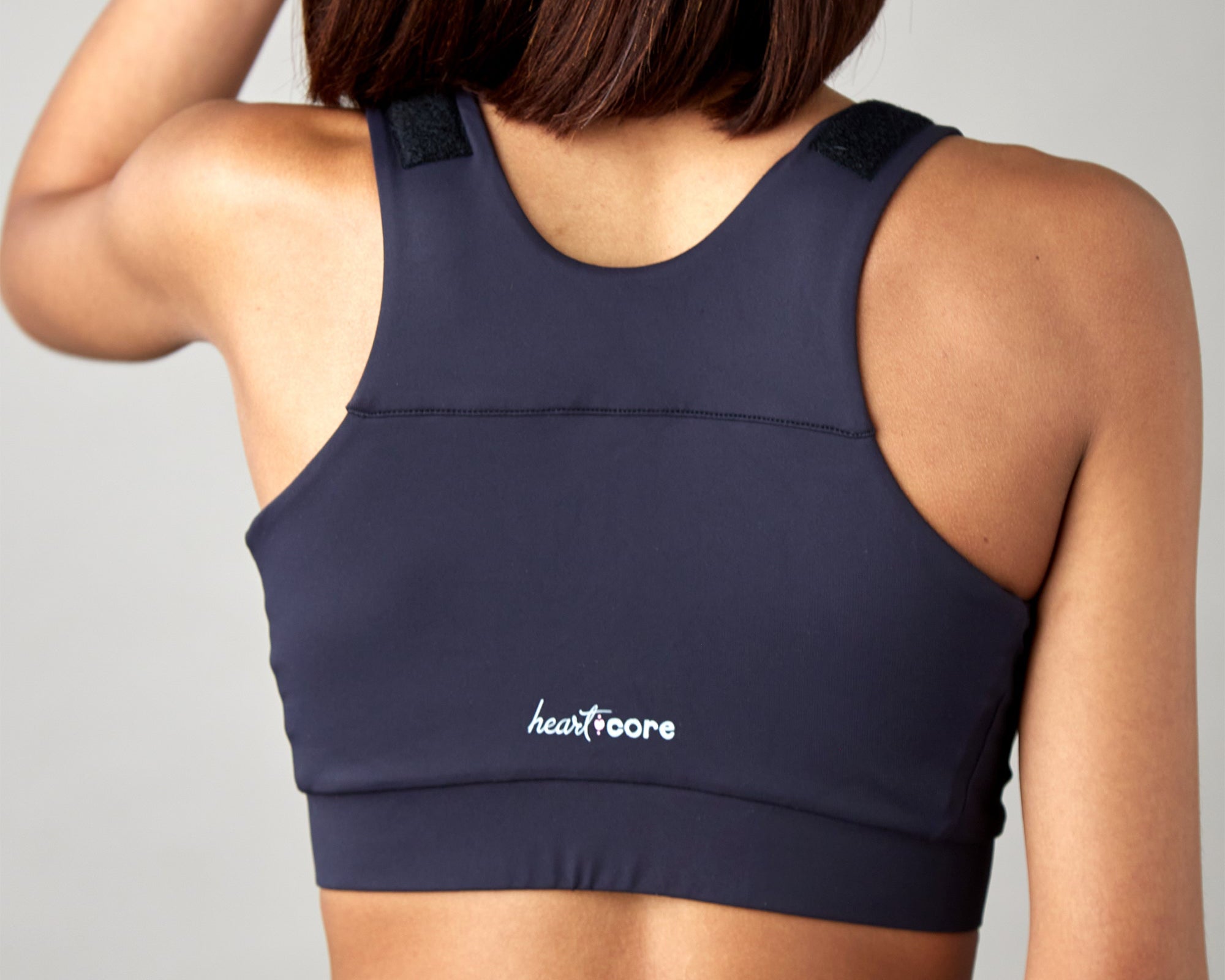 A woman with shoulder-length brown hair stands against a plain light background, wearing the Shirl Post-Surgical Bra in dark color with "heartcore" printed in white on the lower back.