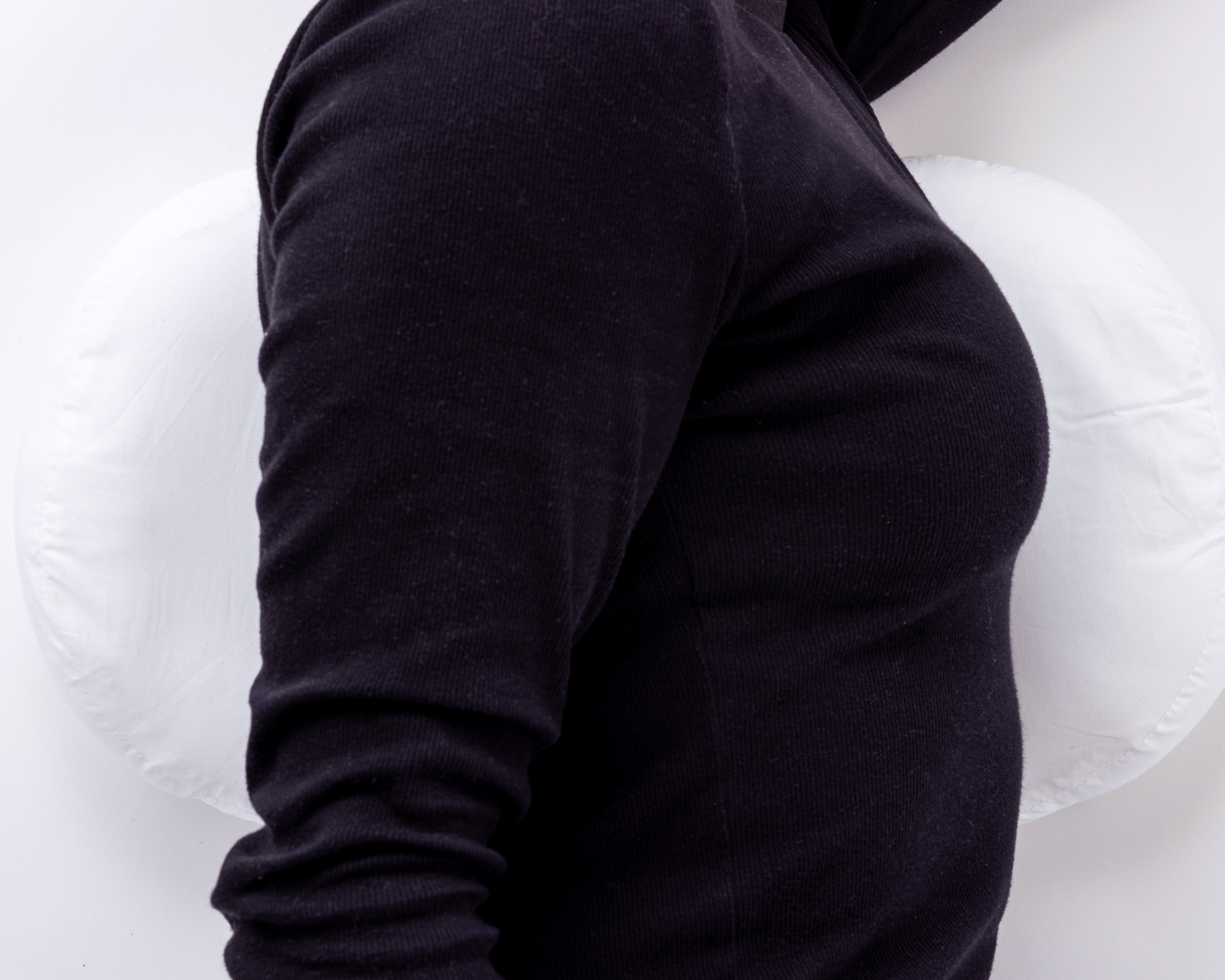 A person in a black long-sleeve shirt uses the Side Sleeping Chest Pillow, sitting with their back against its white, round design for lower back comfort. The background is plain white.
