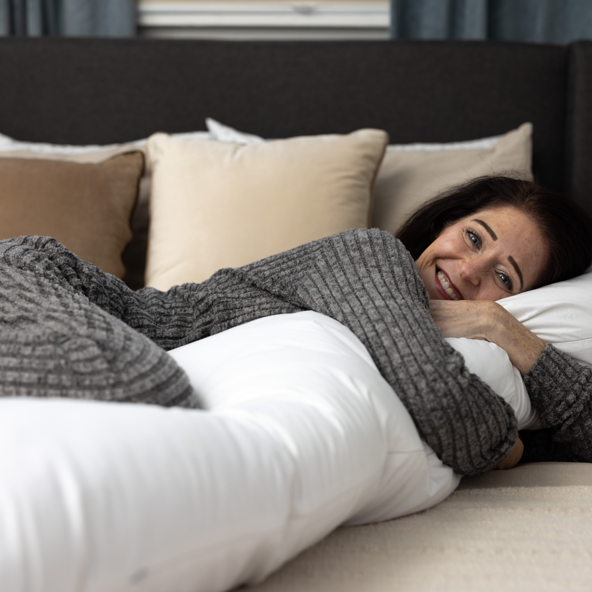 A woman in gray pajamas smiles on a bed, hugging a long white body pillow with a removable washable slipcover. Behind her are beige and white pillows (Two Side Pillows Sold Separately), with blue curtains in the background.
