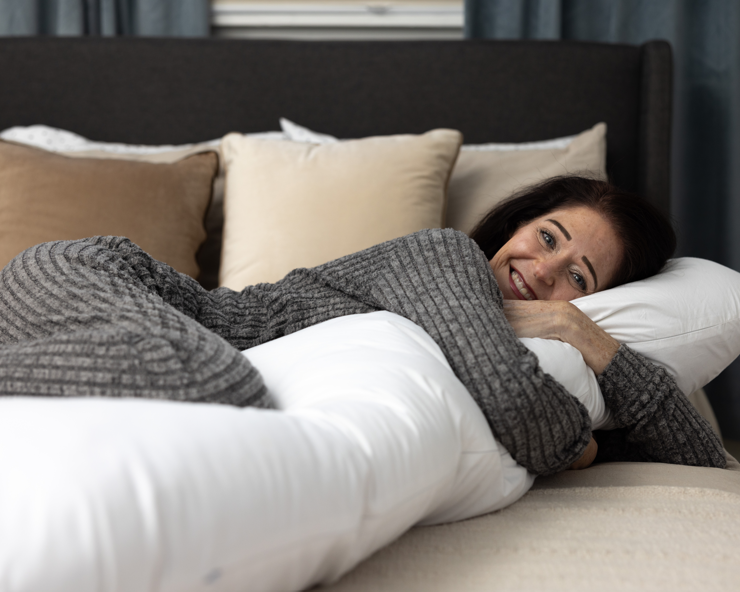 A woman in gray pajamas smiles on a bed, hugging a long white body pillow with a removable washable slipcover. Behind her are beige and white pillows (Two Side Pillows Sold Separately), with blue curtains in the background.