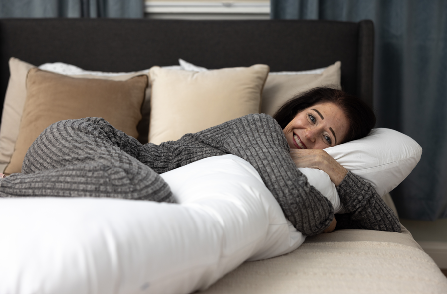 A woman in gray pajamas smiles on a bed, hugging a long white body pillow with a removable washable slipcover. Behind her are beige and white pillows (Two Side Pillows Sold Separately), with blue curtains in the background.