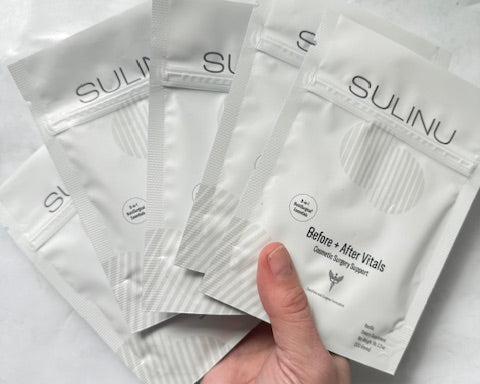 A hand displays five white SULINU Travel Packs—Set of 5, Single Serving Before + After Vitals—vitamin powder, fanned out against a light background. Designed to aid wound healing after surgery and support immunity.