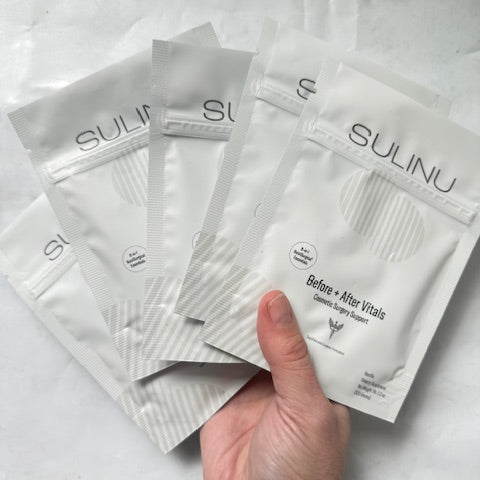 A hand displays five white SULINU Travel Packs—Set of 5, Single Serving Before + After Vitals—vitamin powder, fanned out against a light background. Designed to aid wound healing after surgery and support immunity.