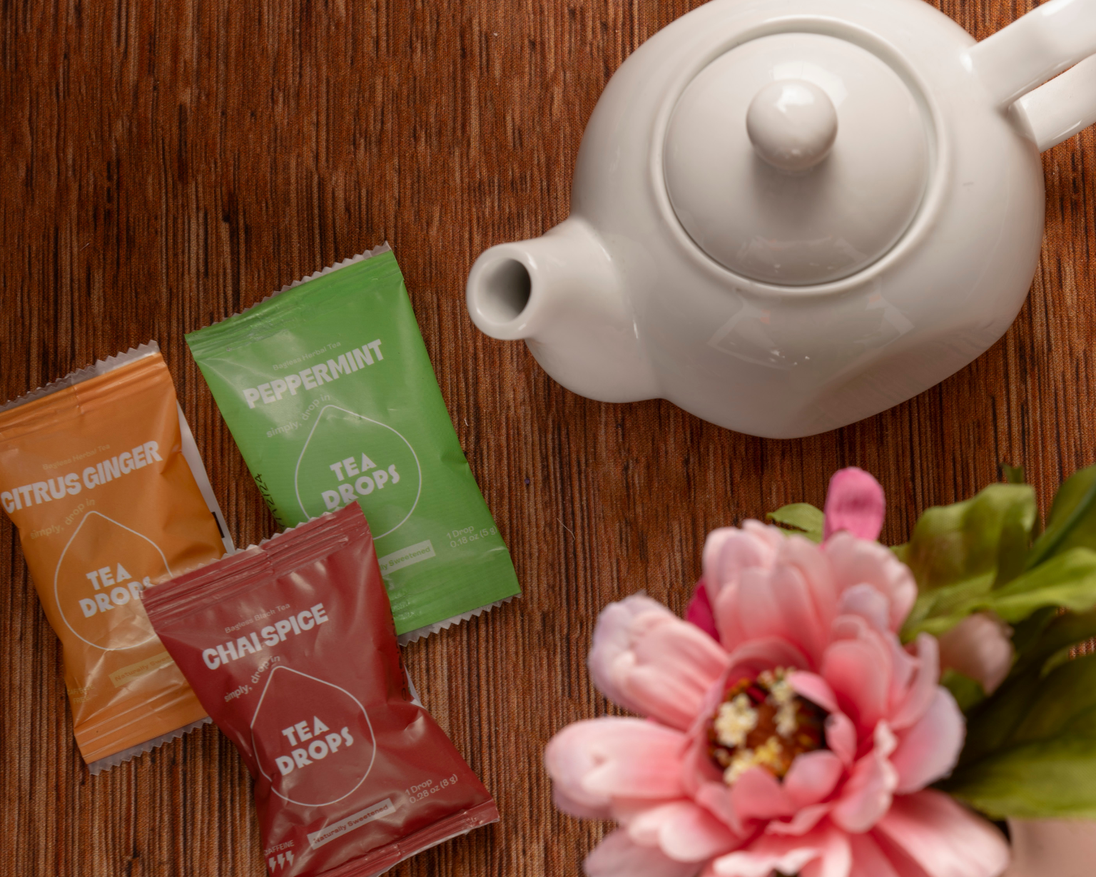 A white teapot sits on wood next to Tea Drops in citrus ginger, peppermint, and chai spice from the Sweetest Rest & Recovery Box—ideal for self-care or cancer recovery. A pink artificial flower with green leaves adds a soft finishing touch.
