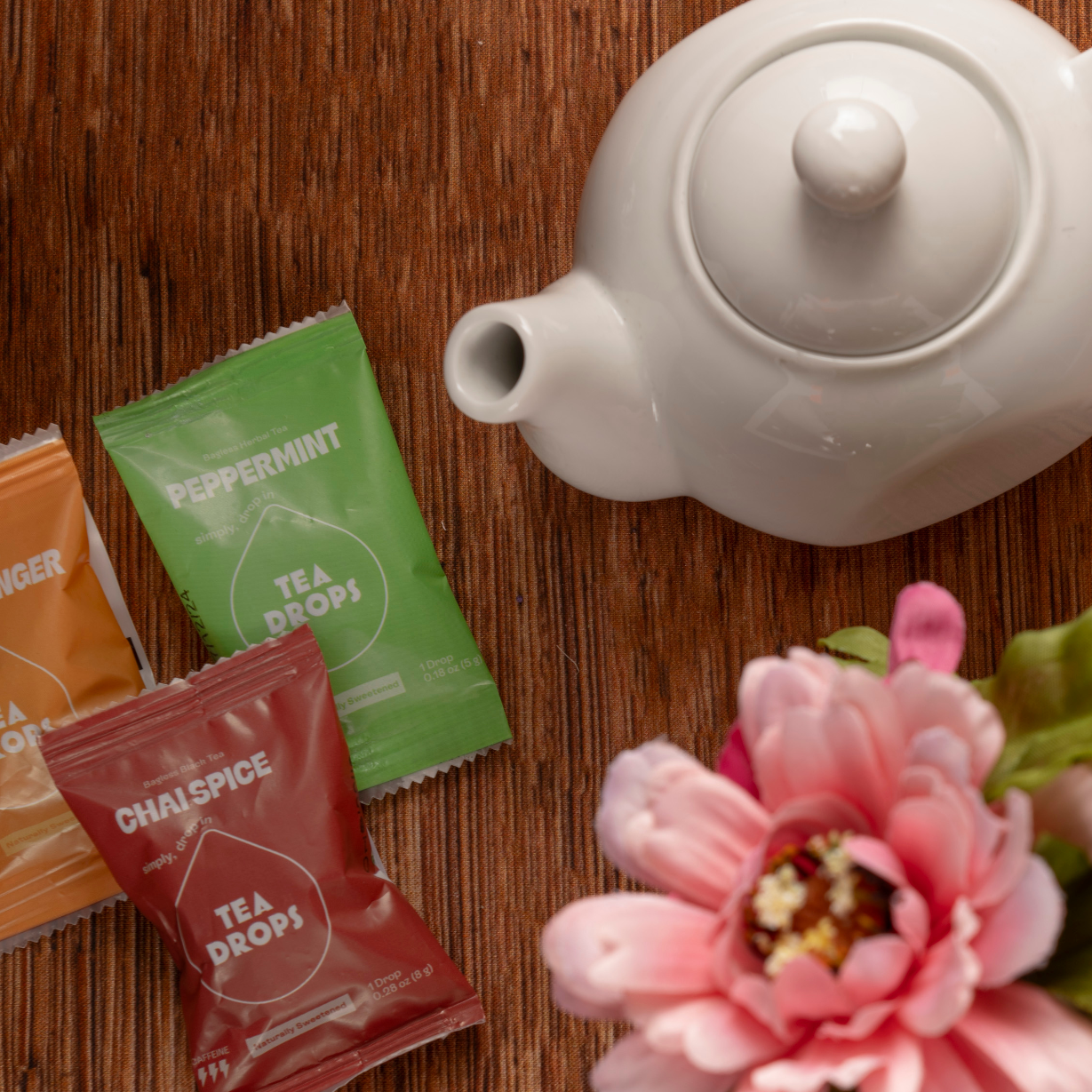 A white teapot sits on wood next to Tea Drops in citrus ginger, peppermint, and chai spice from the Sweetest Rest & Recovery Box—ideal for self-care or cancer recovery. A pink artificial flower with green leaves adds a soft finishing touch.