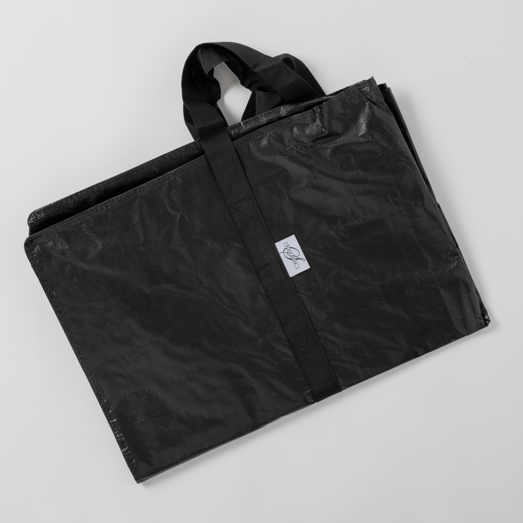 The Travel and Storage Bag for Sleep Again Pillow System is a black, rectangular, water-resistant reusable bag with short handles, folded flat on a white surface. A small white logo label on the front makes it ideal for travel and storage.