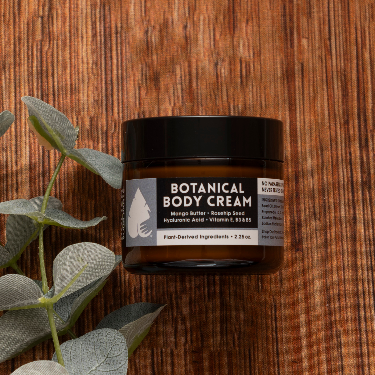 The Best Mastectomy Care Package features a Botanical Body Cream with mango butter, rosehip seed, hyaluronic acid, and vitamin E—perfect for soothing care, beautifully presented beside silver-green eucalyptus on a wooden surface.