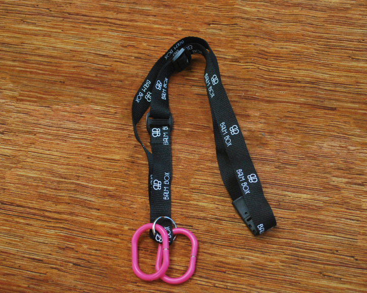 A black “BHA BOX” Surgical Drain Holder lanyard with white text and logo rests on a wooden surface. It features two connected pink plastic clips at one end, making it ideal for use as a mastectomy drain holder.