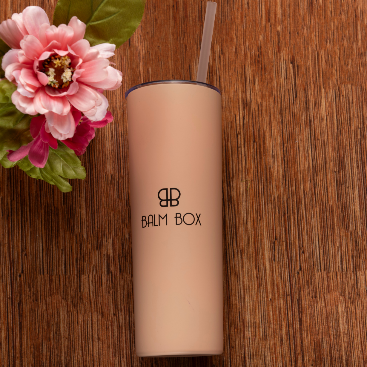 A beige tumbler with straw and BALM BOX logo sits on a wooden surface beside a pink flower—an ideal addition to the Sweetest Rest & Recovery Box, a thoughtful self-care gift for cancer patients.