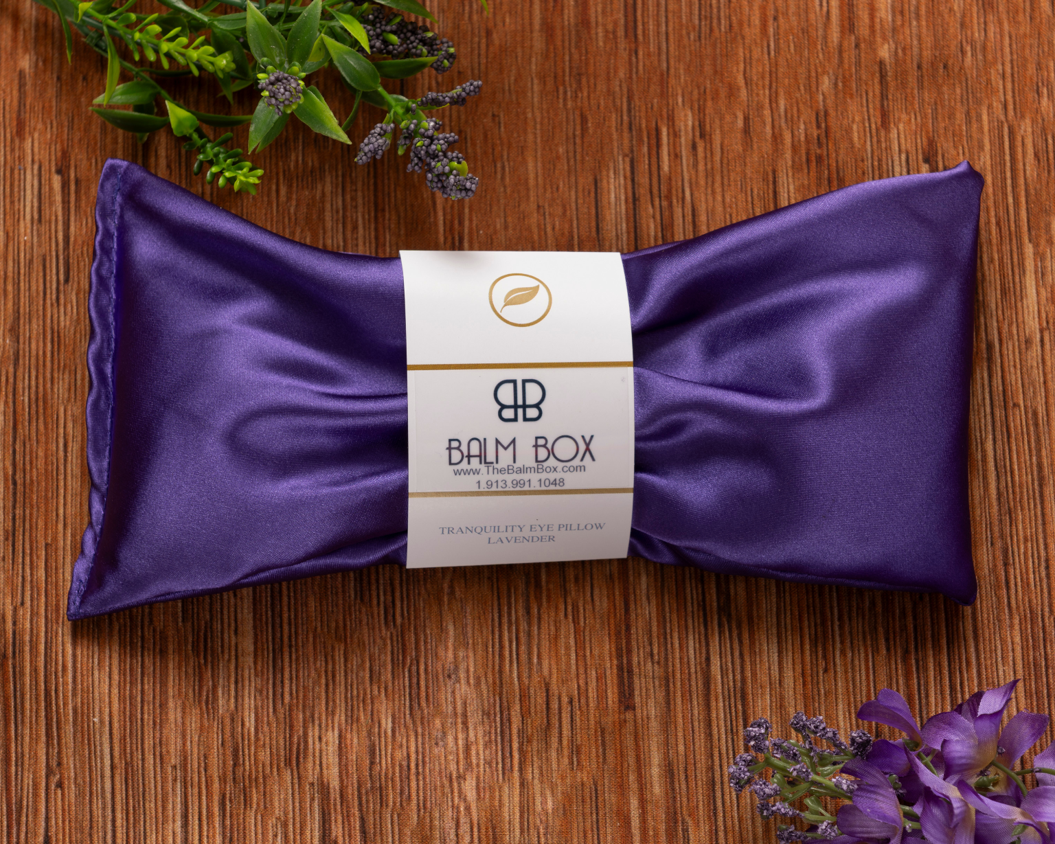 The Sweetest Rest & Recovery Box features a purple silk eye pillow from Balm Box on a wooden surface, surrounded by lavender and greenery—a soothing touch for any cancer recovery care package. The product label is displayed at the pillow’s center.