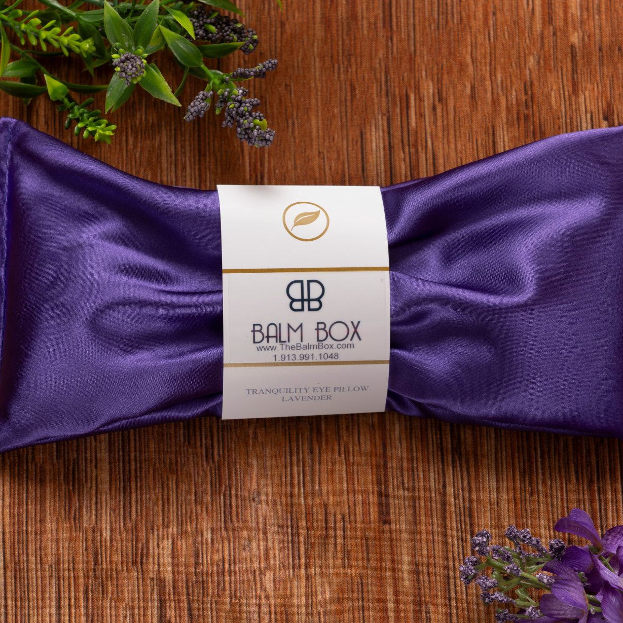 A purple satin eye pillow from The Best Mastectomy Care Package rests on a wooden surface with lavender and green leaves, its Tranquility Eye Pillow Lavender label visible—perfect for a thoughtful breast cancer gift box.