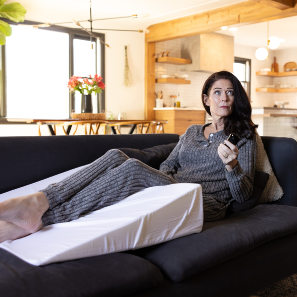 A woman in gray loungewear lounges on a modern couch, feet propped on the Individual Wedge Pillow Sold Separately, as she holds a remote and looks aside in a bright living room.