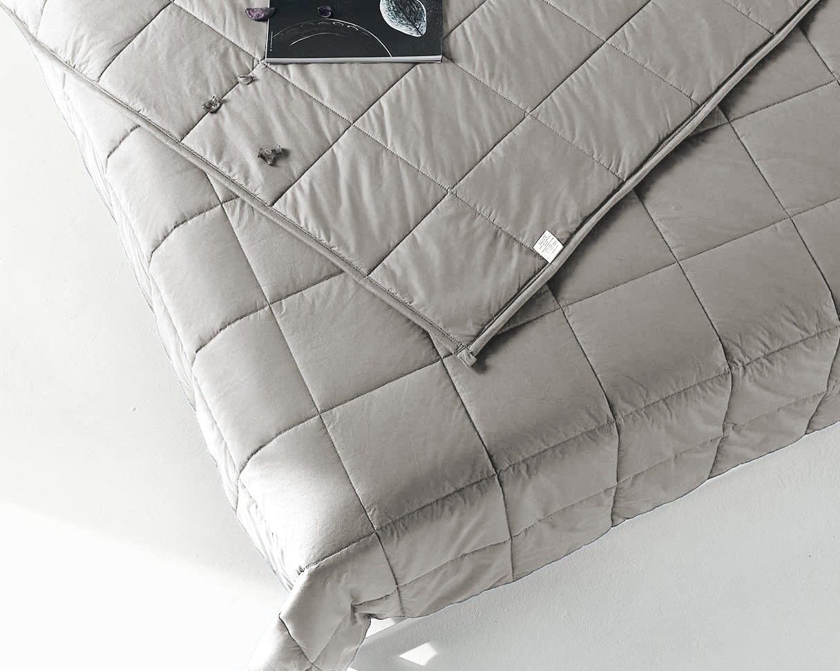 A minimal, modern bed features the Pillowtex Weighted Blanket in light grey, quilted and filled with glass beads. An open book and eyeglasses rest on the corner of the neatly made bed.