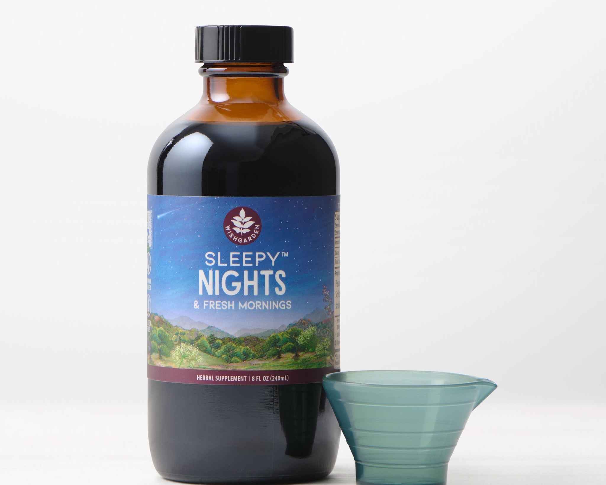 A brown bottle of Sleepy Nights & Fresh Mornings organic sleep supplement, featuring a night sky and green landscape on the label, stands beside a small blue plastic dosage cup on a white surface.