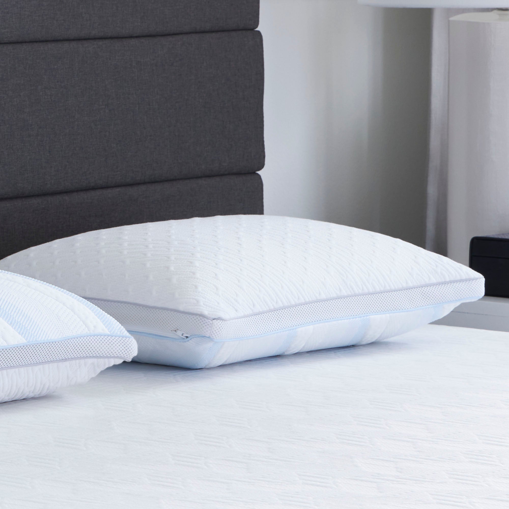 Two Comfort Tech Dual Comfort Foam Pillows with textured white covers rest on a neatly made bed with a white sheet, while a dark gray upholstered headboard and part of a nightstand with a lamp are visible in the background.